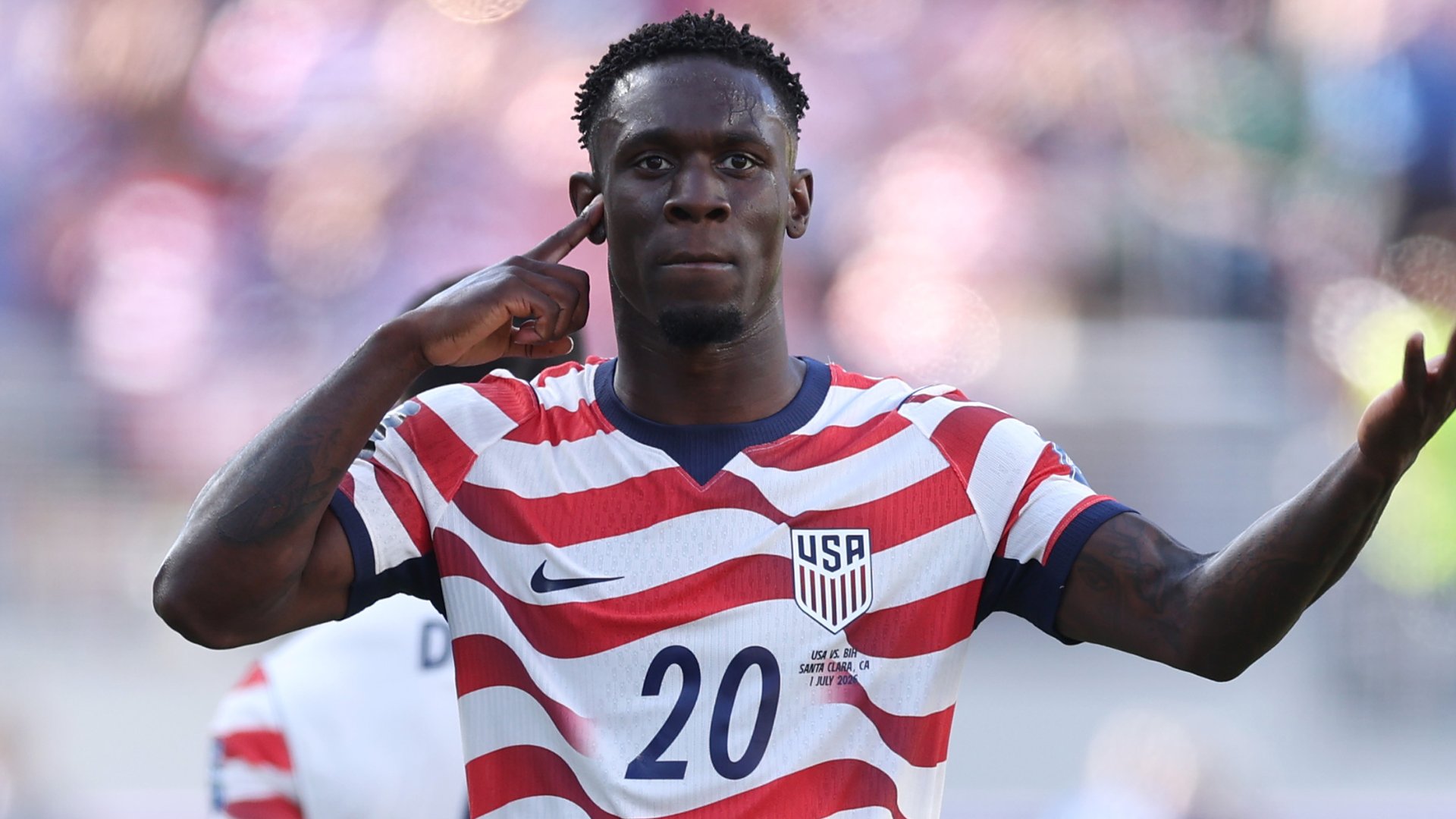 A soccer player wearing a USA #20 red, white, and blue striped jersey points to his ear while gesturing during a match, with text reading "USA vs. BIH, Santa Clara, CA, July 2025."