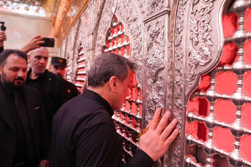 A man presses his hand against an ornate silver grille with red backing, surrounded by others in dark clothing and uniformed personnel observing in an elaborately decorated interior.