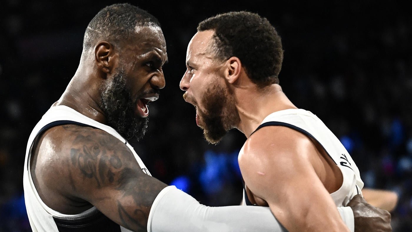 Two bearded basketball players in white jerseys stand face-to-face, shouting at each other during an indoor game.