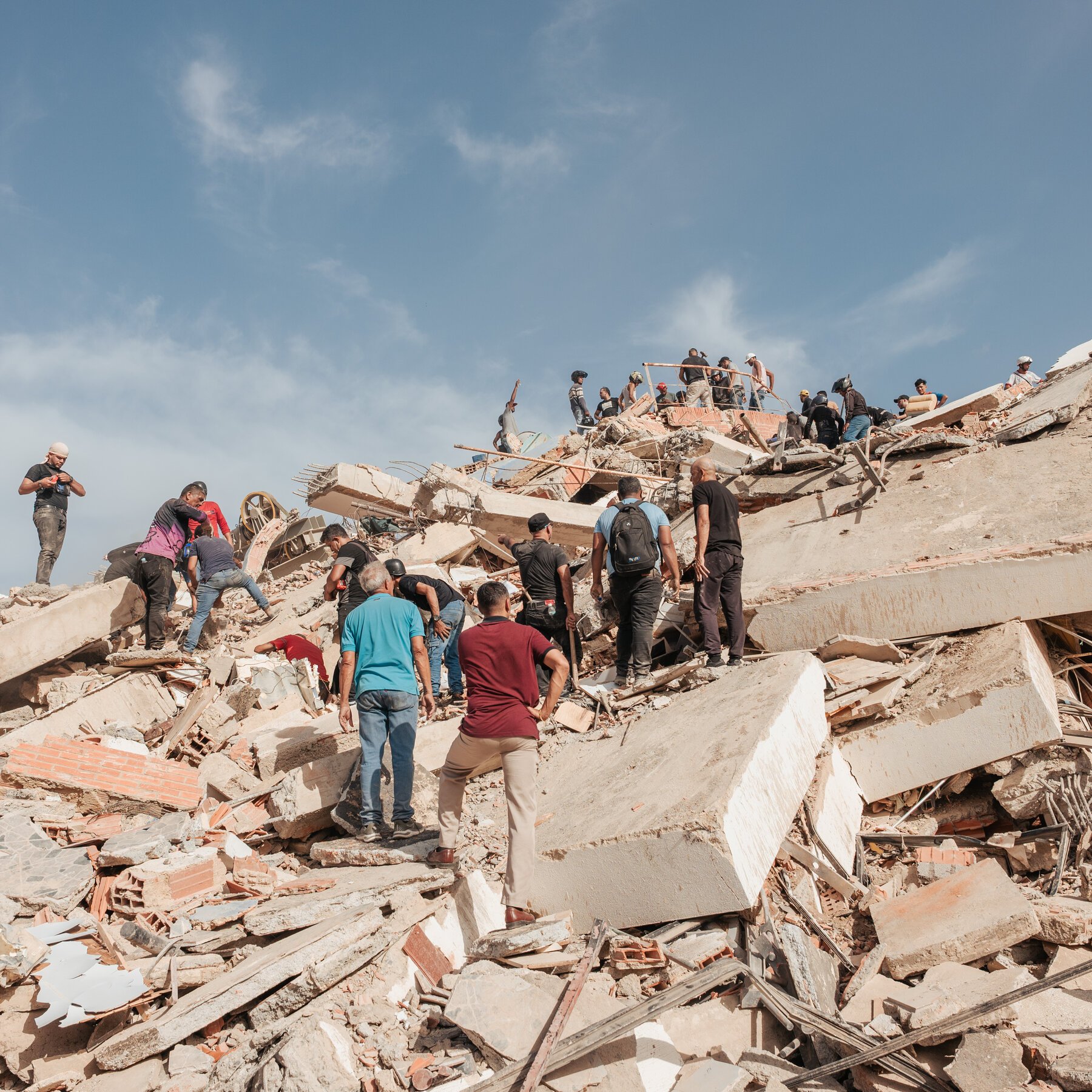 Numerous people climb and search through a large pile of concrete and brick rubble under a blue sky.
