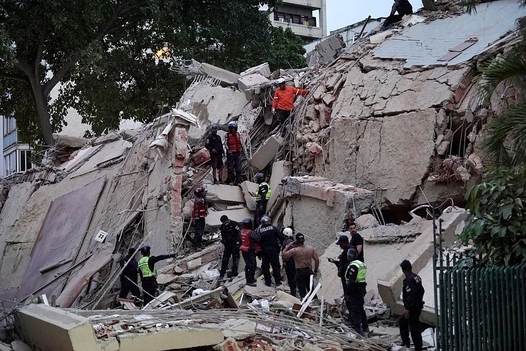 Rescue workers in helmets and uniforms climb atop a massive pile of concrete rubble and twisted rebar from a collapsed building, conducting search operations amid scattered debris.