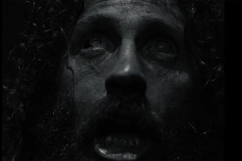 A close-up, black-and-white image shows a bearded man's face with dark, sunken eyes and a tear-streaked cheek, his mouth slightly open.