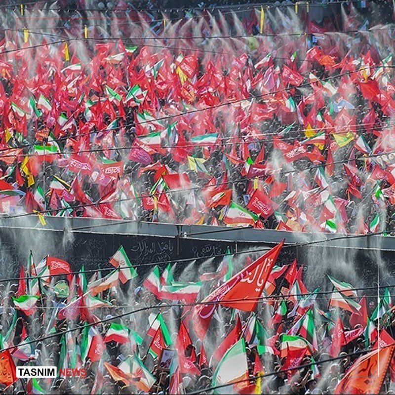 A large crowd waves numerous red, green, and white flags amid heavy spray or smoke during a public gathering, with Arabic-script banners visible overhead.