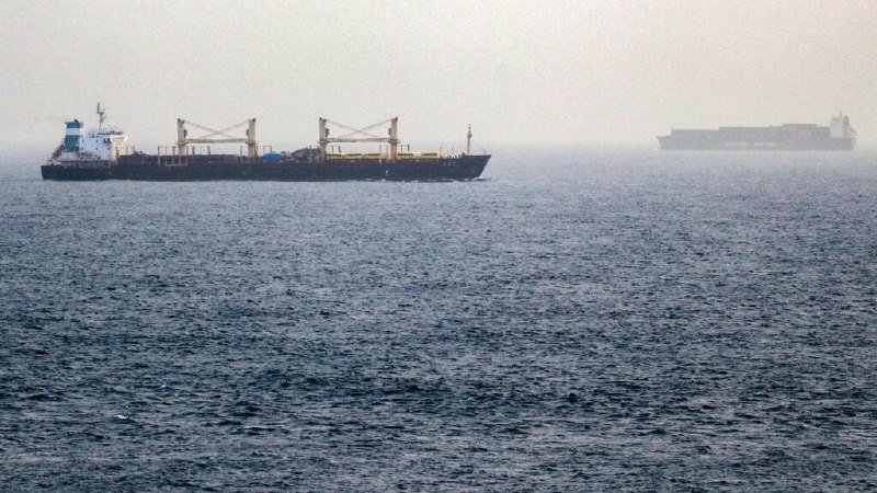 A large cargo ship with cranes sails on the open water, with another vessel faintly visible in the hazy distance.