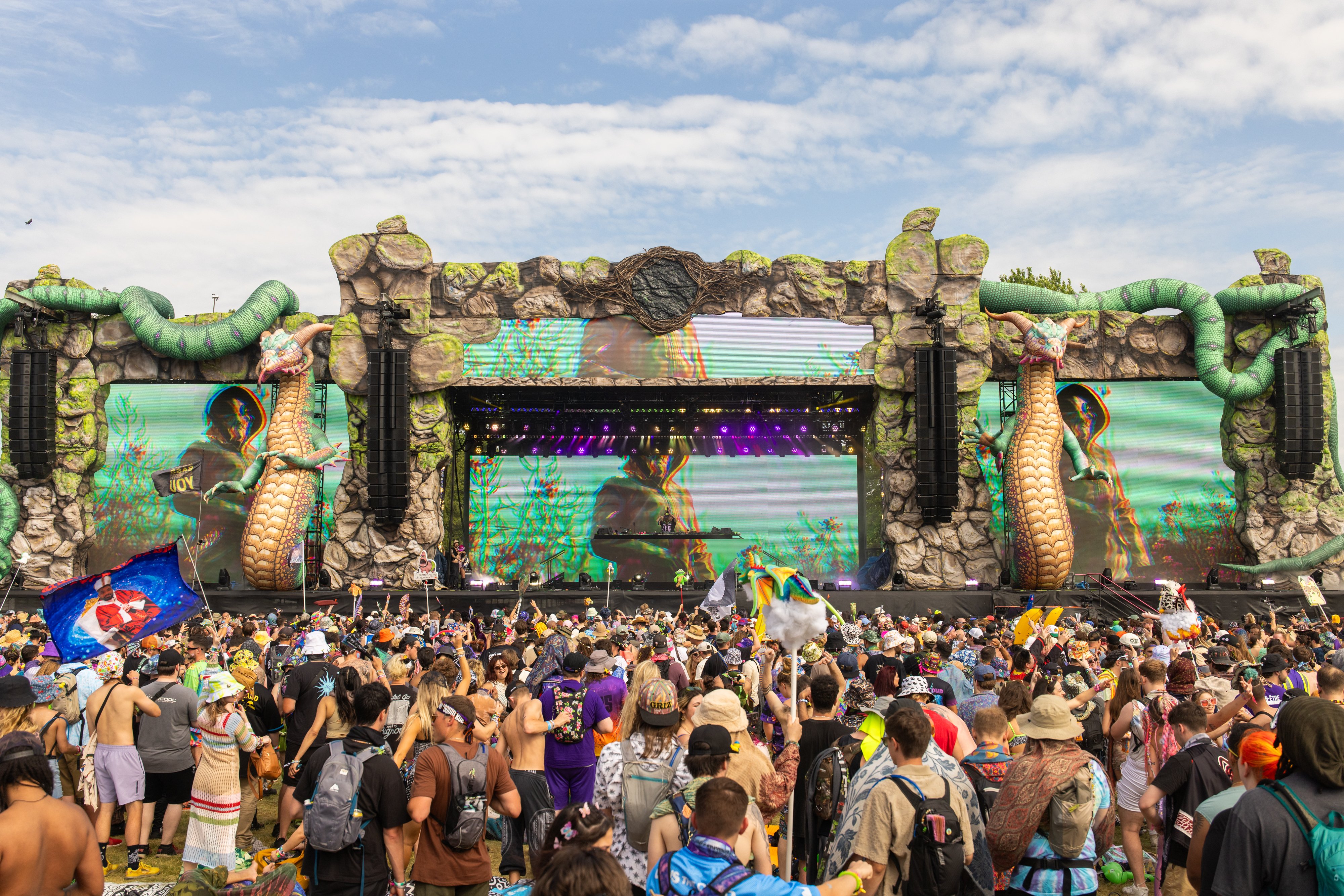 A large crowd of festivalgoers gathers before an elaborate outdoor stage featuring rock-like structures, giant green serpent sculptures, and large LED screens displaying colorful visuals under a partly cloudy sky.