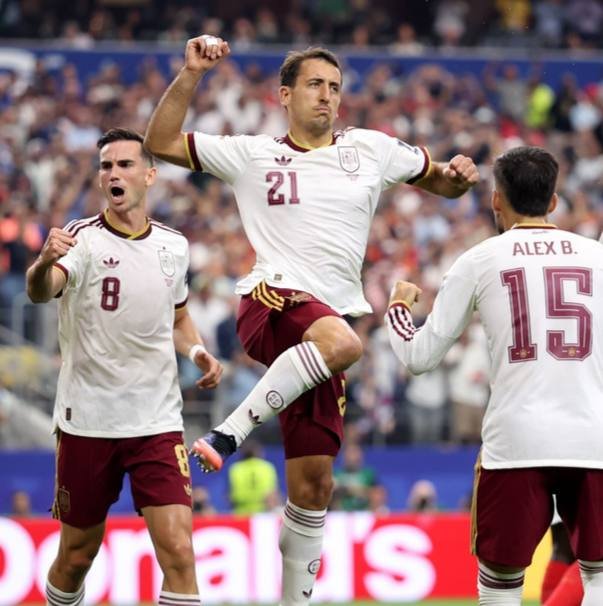 Three soccer players in white and maroon jerseys celebrate on the field, with the player wearing number 21 leaping with a raised fist while teammates numbered 8 and "ALEX B. 15" shout nearby.