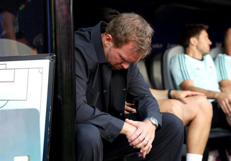 A man in a dark suit sits on a bench with his head bowed and hands clasped, while two men in light blue athletic shirts sit behind him.