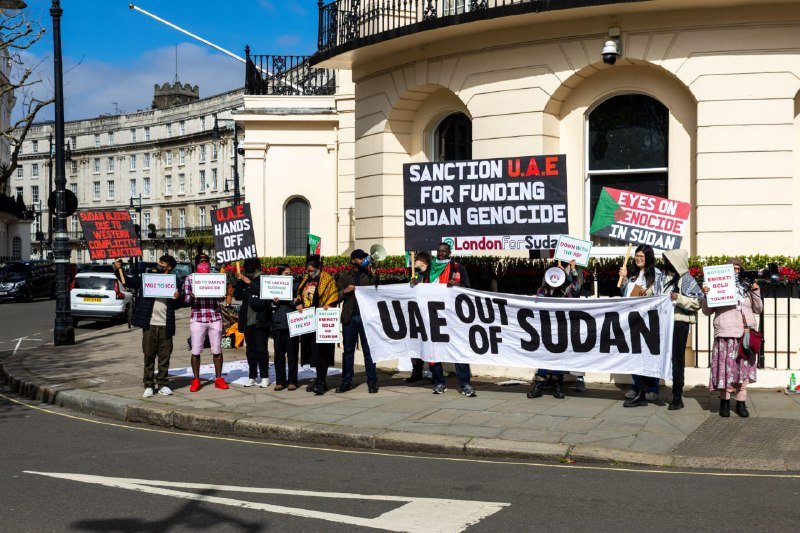 Protesters hold signs and a banner reading "UAE OUT OF SUDAN" and "SANCTION U.A.E FOR FUNDING SUDAN GENOCIDE" outside a cream-colored building on a city street.