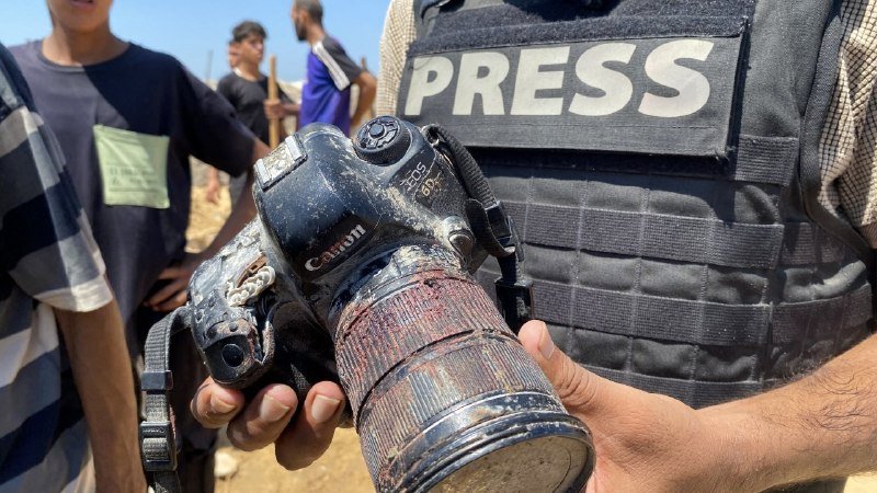 A person wearing a "PRESS" vest holds a mud-splattered Canon EOS DSLR camera outdoors near other individuals.