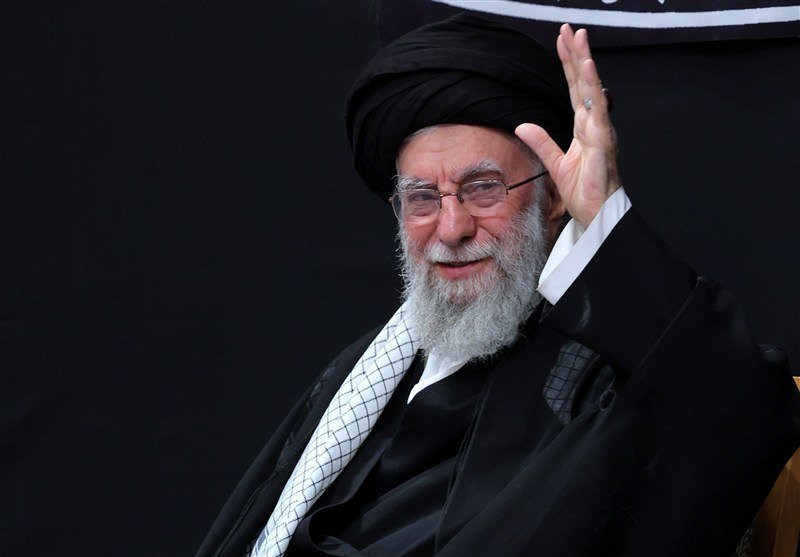 A bearded man wearing a black turban and robes, with glasses and a checkered scarf, smiles and raises one hand against a dark background.