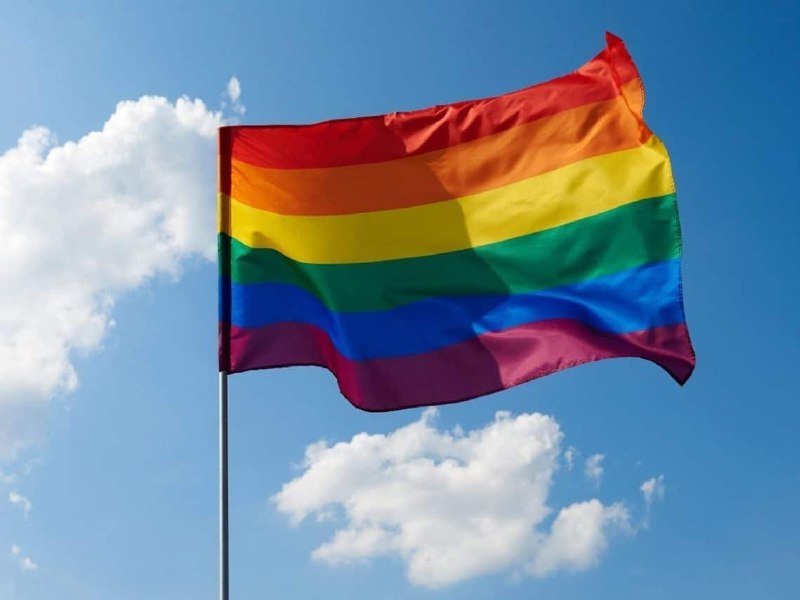A rainbow-striped flag waves on a pole against a blue sky with scattered white clouds.