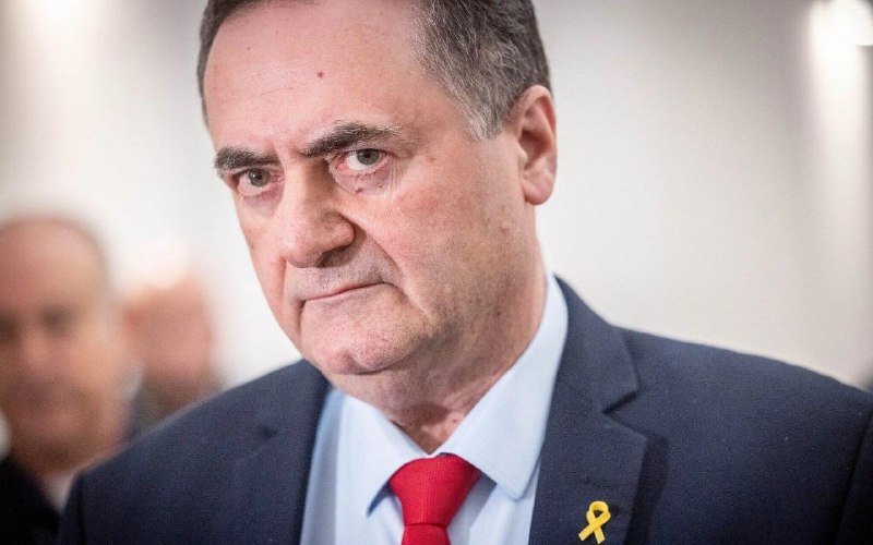 A serious-looking man in a dark suit, light blue shirt, and red tie with a yellow ribbon pin, photographed in a close-up portrait with a blurred background.