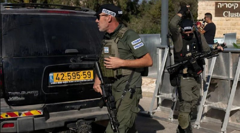 Armed police officers in olive-green uniforms with "POLICE" headbands and helmets stand near a black Chevrolet Trailblazer near a metal barricade, with one aiming a rifle.