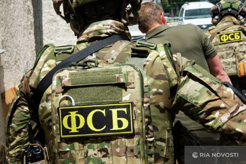 Armed personnel in camouflage gear escorting a man in a green shirt, with vests displaying "ФСБ" patches, on a street beside a white vehicle.