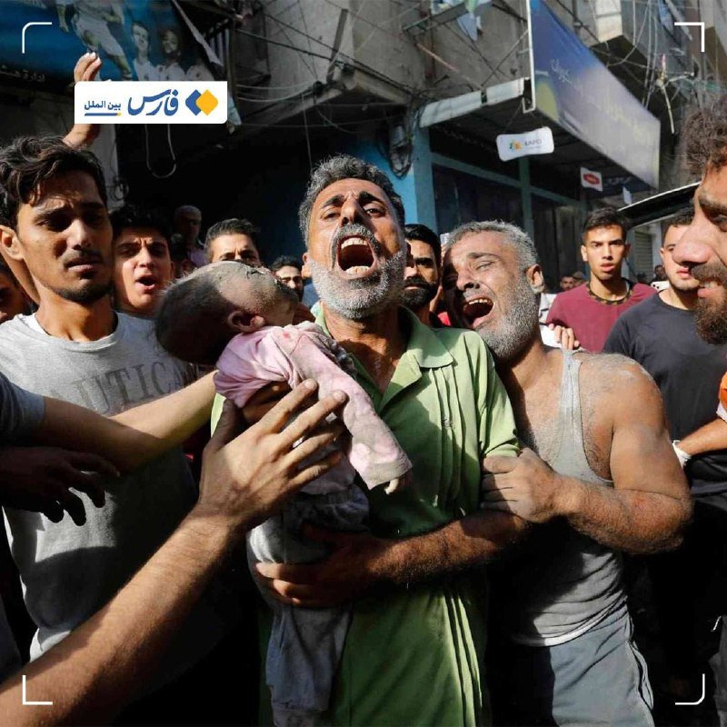 A grieving man cries out while holding a small child, surrounded by a crowd of distressed people in a street setting with shops visible in the background.