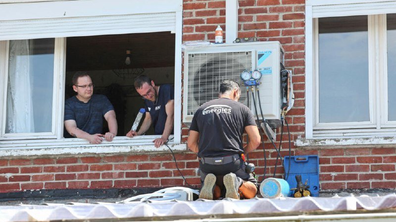 A technician in an "Energies" shirt kneels installing an air conditioning unit on a brick wall, observed by two men through a window.