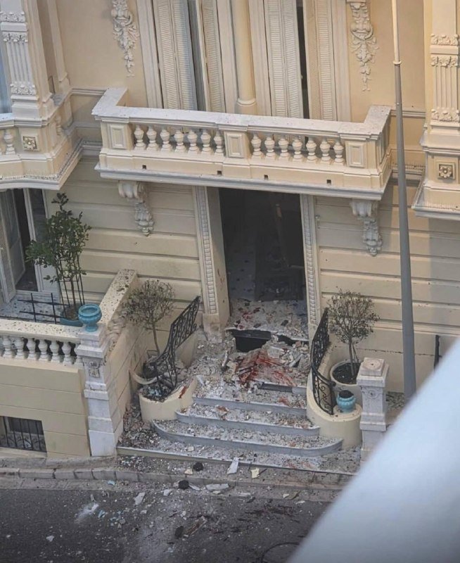 Debris and shattered stone cover the entrance steps of an ornate cream-colored building, with broken fragments scattered across the doorway and sidewalk below.
