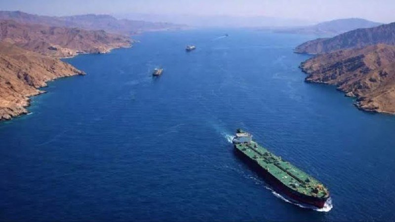 Aerial view of a large green-hulled cargo ship traveling through a narrow strait flanked by arid, mountainous coastlines, with smaller vessels visible in the distance.