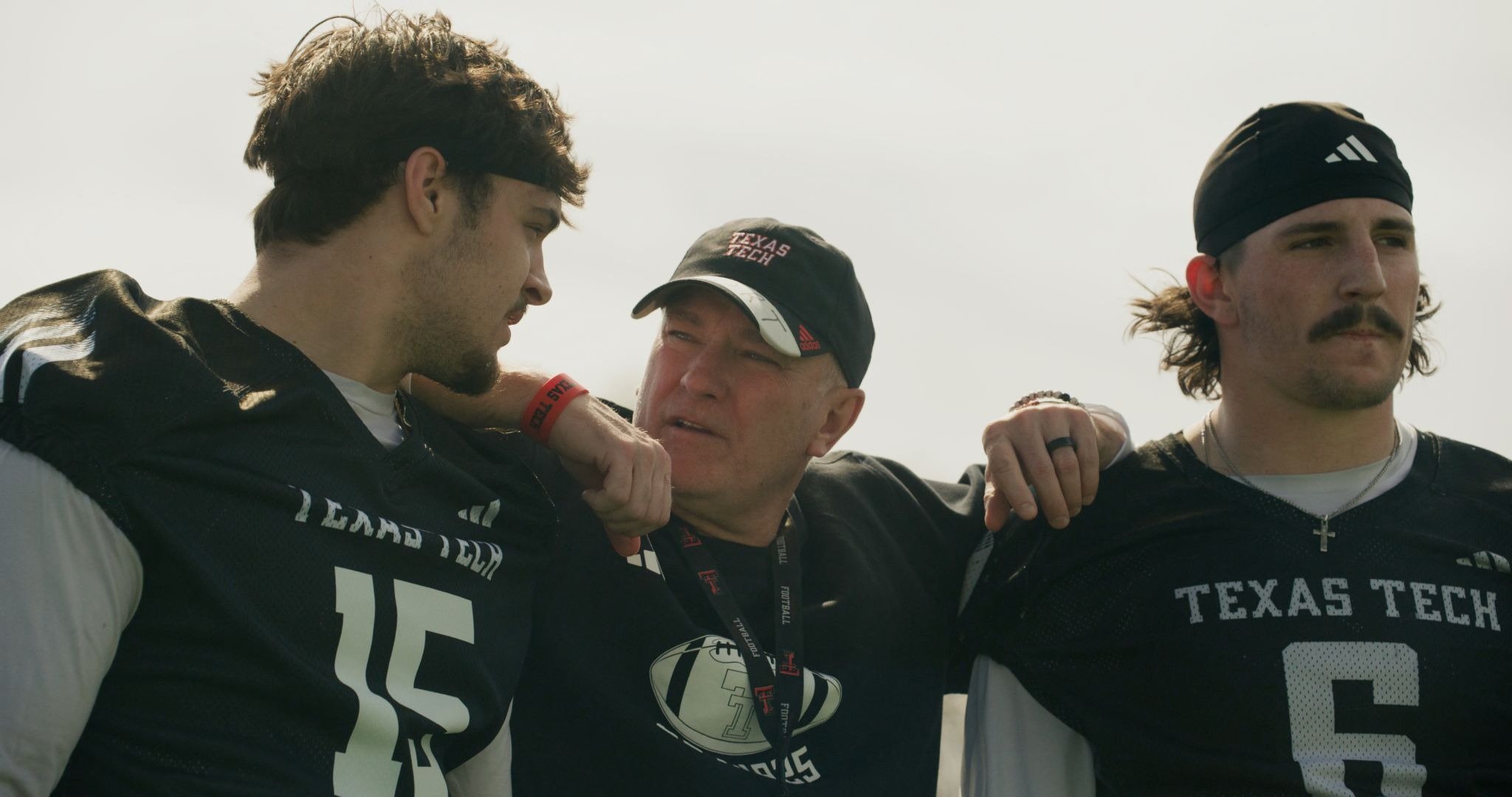 Three men in Texas Tech football jerseys stand together outdoors, with the center man wearing a "Texas Tech" cap and his arms around the two younger players on either side.
