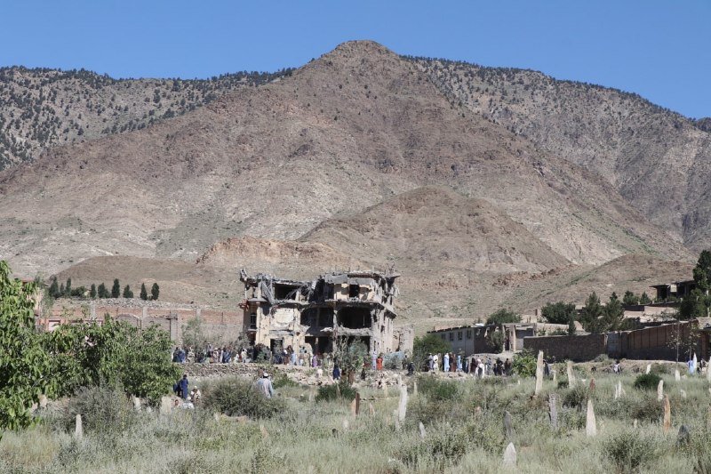A heavily damaged multi-story building stands among a grassy area with scattered grave markers, set against a backdrop of arid, rocky mountains under a clear blue sky.