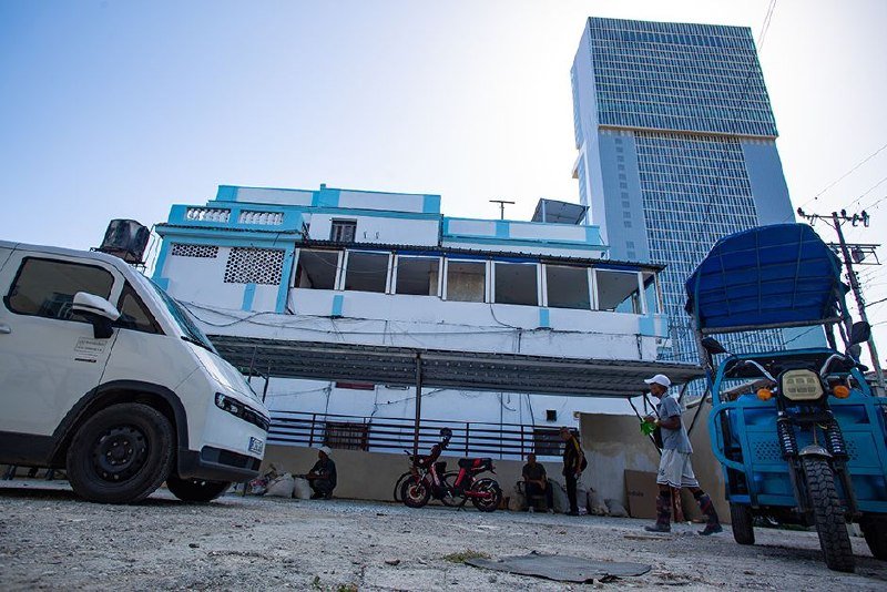 A white van, red motorcycle, and blue auto-rickshaw are parked on a gravel lot in front of a white building and a tall glass skyscraper.