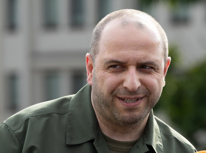 A balding man with a light beard smiles while wearing an olive green shirt, with blurred buildings and trees visible in the background.