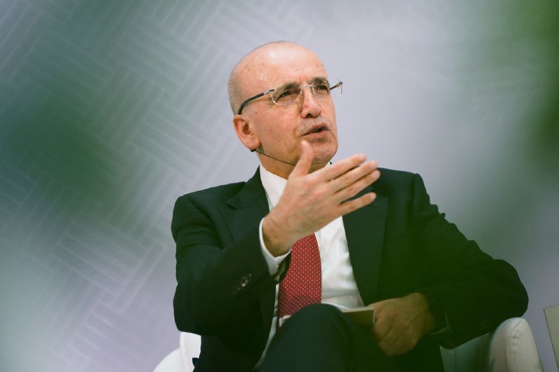 A bald man wearing glasses, a dark suit, and red tie speaks while gesturing with his hand, seated against a blurred gray-green background.