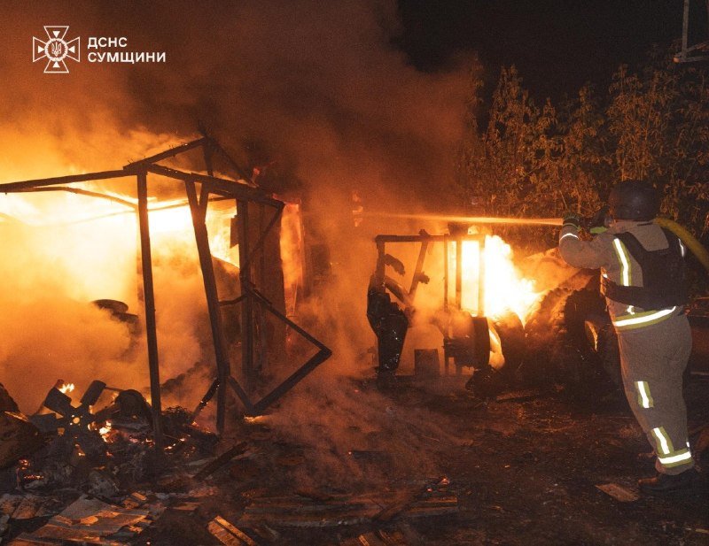 A firefighter in reflective gear sprays water from a hose at a large nighttime fire engulfing a collapsed structure, with a logo reading "ДСНС СУМЩИНИ" in the corner.