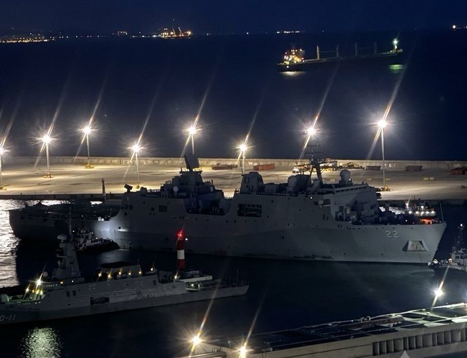 A large gray naval warship marked "22" is docked at a brightly lit harbor at night, with smaller vessels and a cargo ship visible in the background.