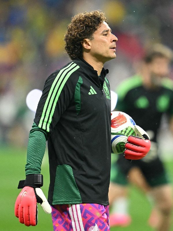 A curly-haired goalkeeper in a black and green Mexico training kit and pink gloves holds a soccer ball during what appears to be a warm-up session on the pitch.
