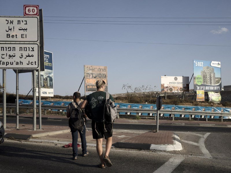 Two backpackers stand on a roadside near Hebrew road signs for Bet El and Tapu'ah Junction, with billboards visible in the background.