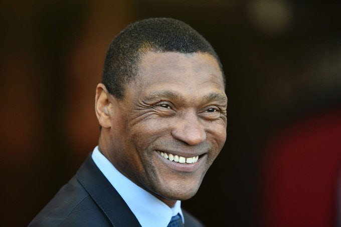 A smiling man in a dark suit, light blue shirt, and dark tie poses for a photo against a dark background.