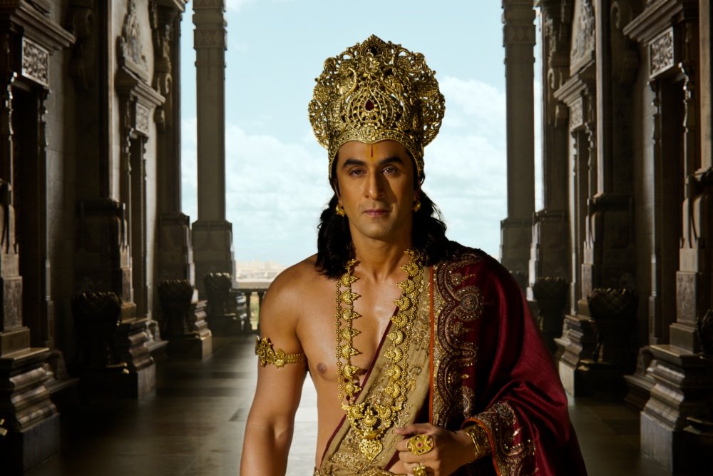 A man with long dark hair wearing an ornate golden crown, gold necklaces, and a red and gold robe stands shirtless between carved stone pillars.