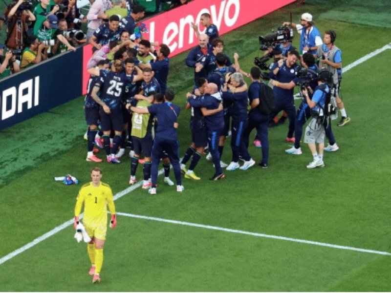 A team in dark blue jerseys celebrates in a group huddle on a grass field near a "Lenovo" advertising board, while a lone goalkeeper in yellow walks away holding a water bottle.