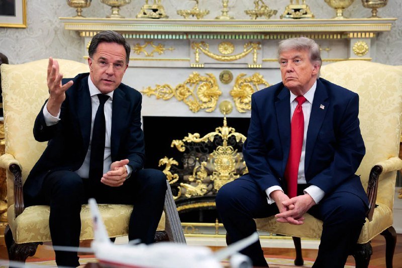Two men in dark suits sit in ornate yellow chairs before a gilded fireplace, one gesturing while speaking and the other listening with hands clasped.