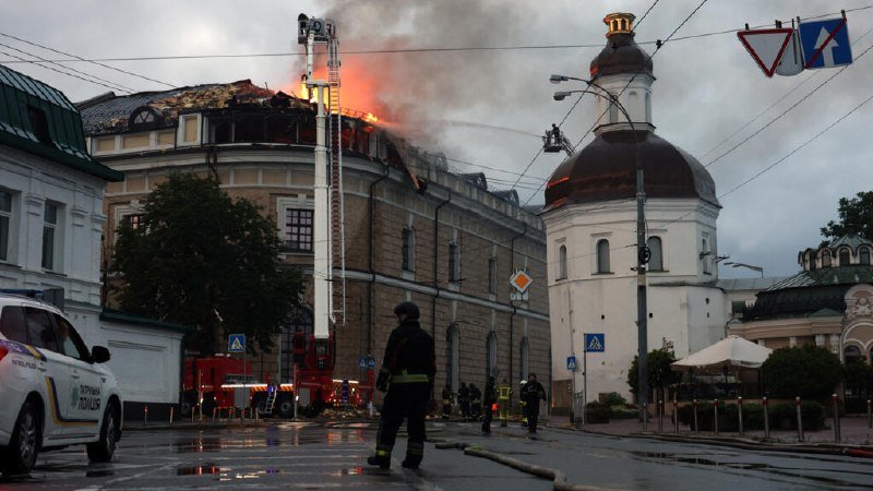 Flames and heavy smoke pour from the roof of an ornate multi-story building as firefighters work below on a wet city street.