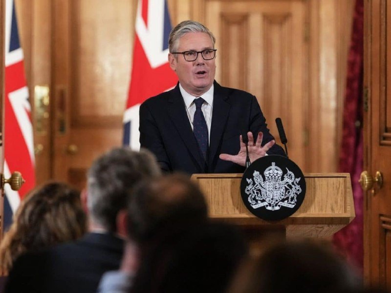 A gray-haired man in a dark suit gestures while speaking at a wooden podium displaying a royal crest, with Union Jack flags and a seated audience visible.