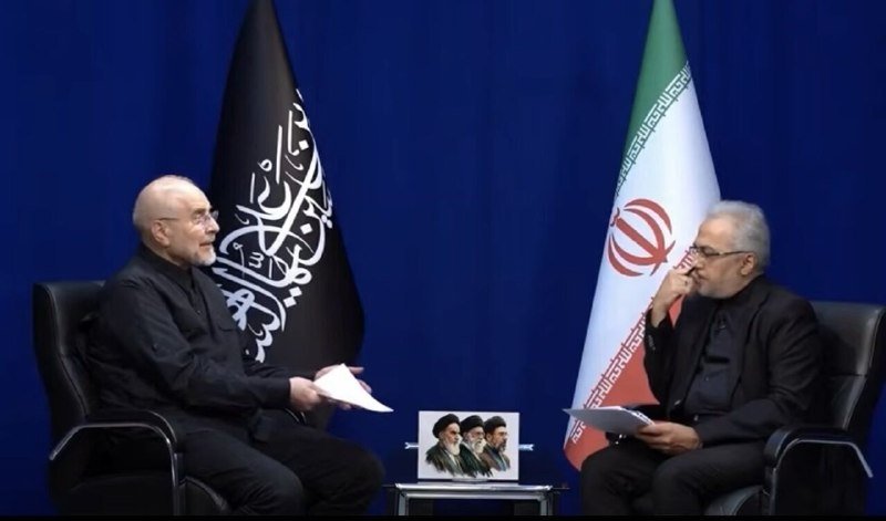 Two men in dark clothing sit facing each other in armchairs against a blue backdrop displaying black and green-white-red flags.