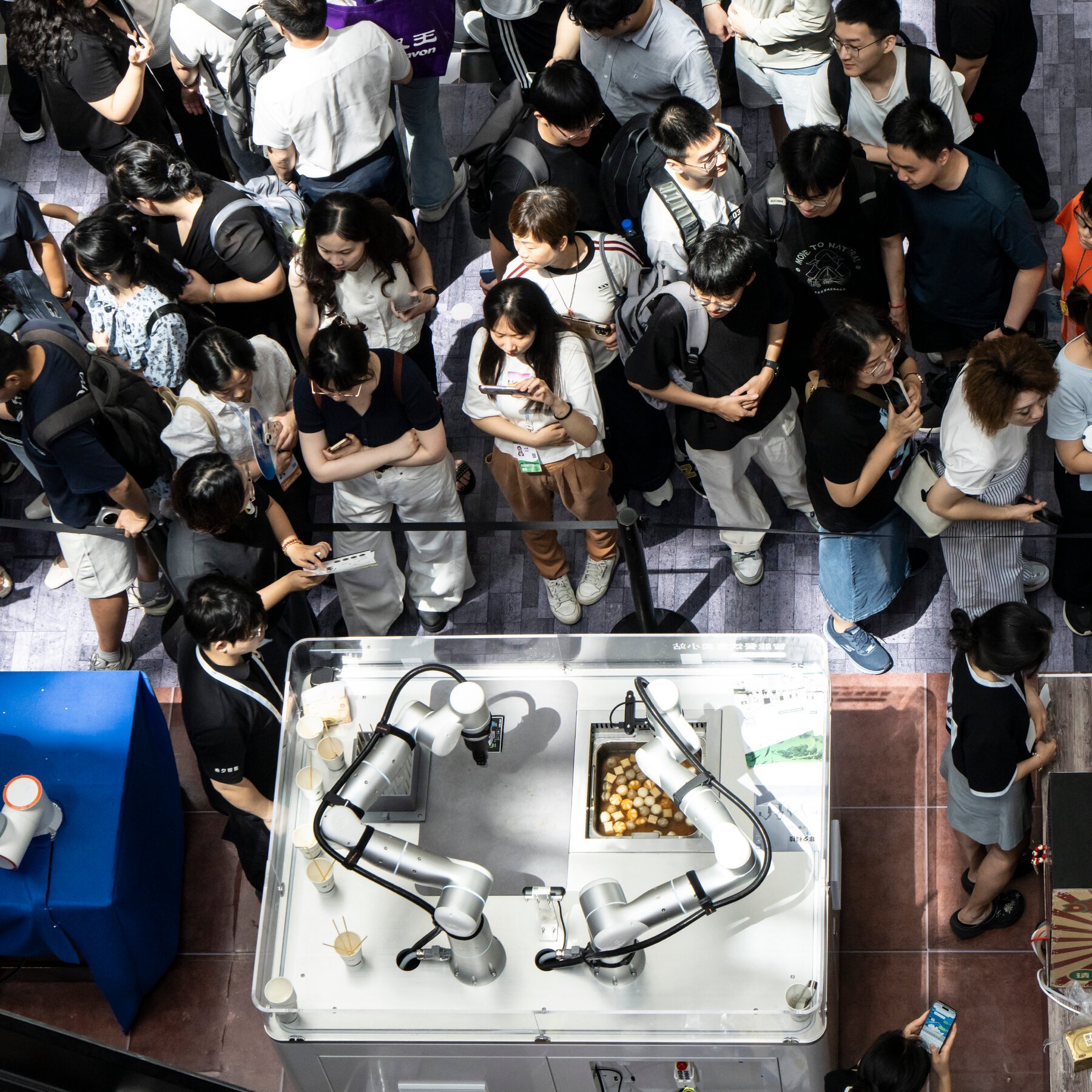 A crowd of people looks down at a robotic arm inside a clear display case, with several individuals holding smartphones to record or photograph the demonstration.