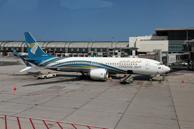 An Oman Air passenger jet is parked on the tarmac at an airport terminal, with ground service equipment positioned nearby under a clear blue sky.