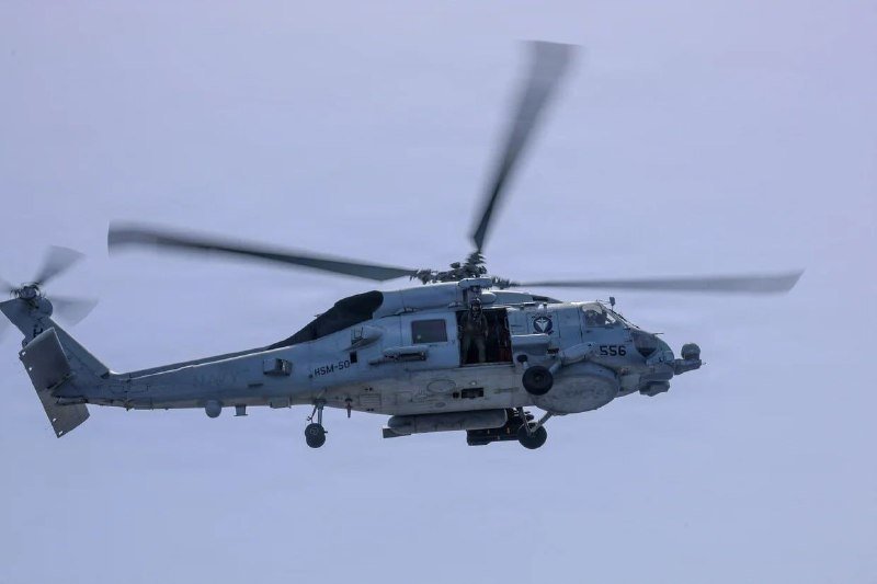 A gray military helicopter marked "556" and "HSM-50" flies against a pale sky, its rotor blades spinning with a crew member visible in the open side door.