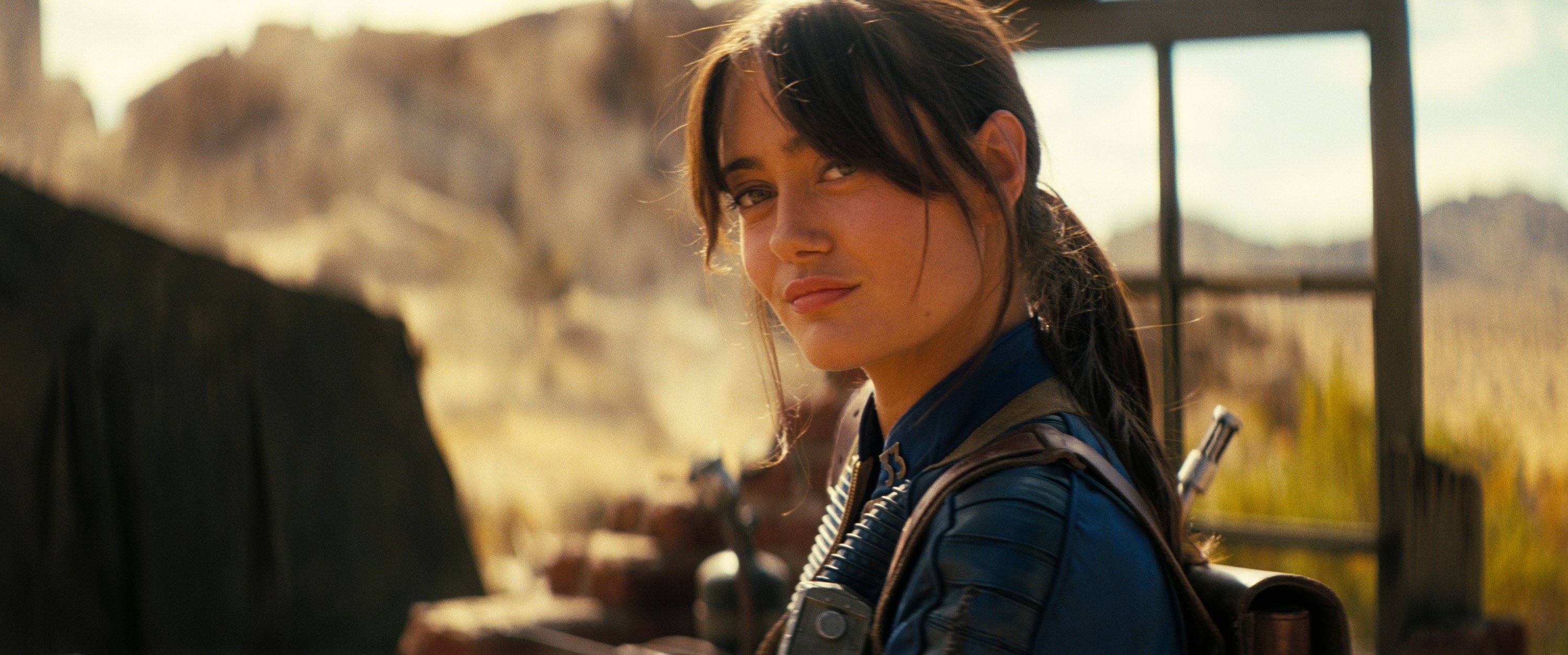A woman in a blue uniform with a ponytail looks over her shoulder at the camera, set against a mountainous backdrop.