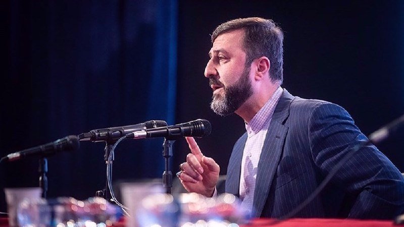 A bearded man in a dark pinstripe suit speaks into a microphone, gesturing with his index finger raised, at a table with a red tablecloth.