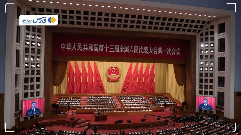 A large assembly hall features a stage with a national emblem, red drapery, and Chinese characters, while attendees sit at desks below and a man appears on a screen at the podium.