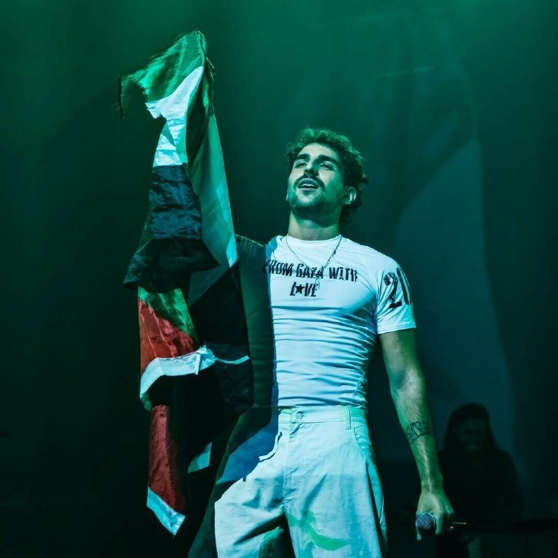 A bearded man in a white graphic t-shirt and light pants waves a red, white, and blue flag on a dimly lit stage bathed in green light.