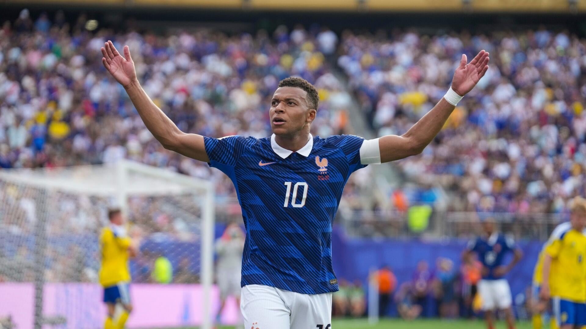 A soccer player in a blue number 10 jersey stands with arms raised in celebration on a stadium pitch before a packed crowd.
