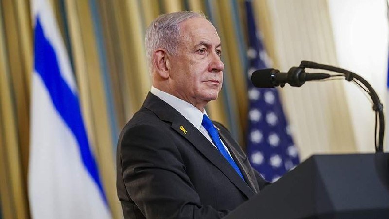 A gray-haired man in a dark suit with a yellow ribbon pin stands at a podium, speaking into a microphone, with Israeli and American flags visible behind him.