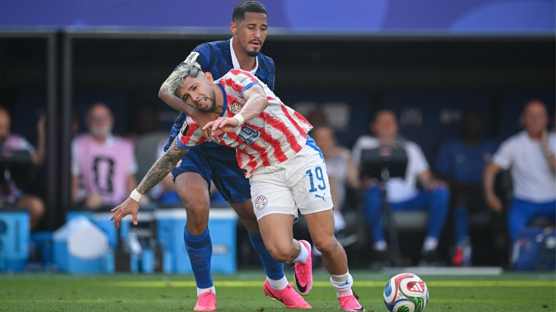 Two soccer players compete for the ball, one in a red-and-white striped #19 jersey, the other in dark blue, with teammates and staff visible on the bench behind them.