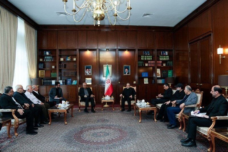 A group of men sit in a wood-paneled meeting room with bookshelves, an Iranian flag, and two framed portraits displayed on the back wall.