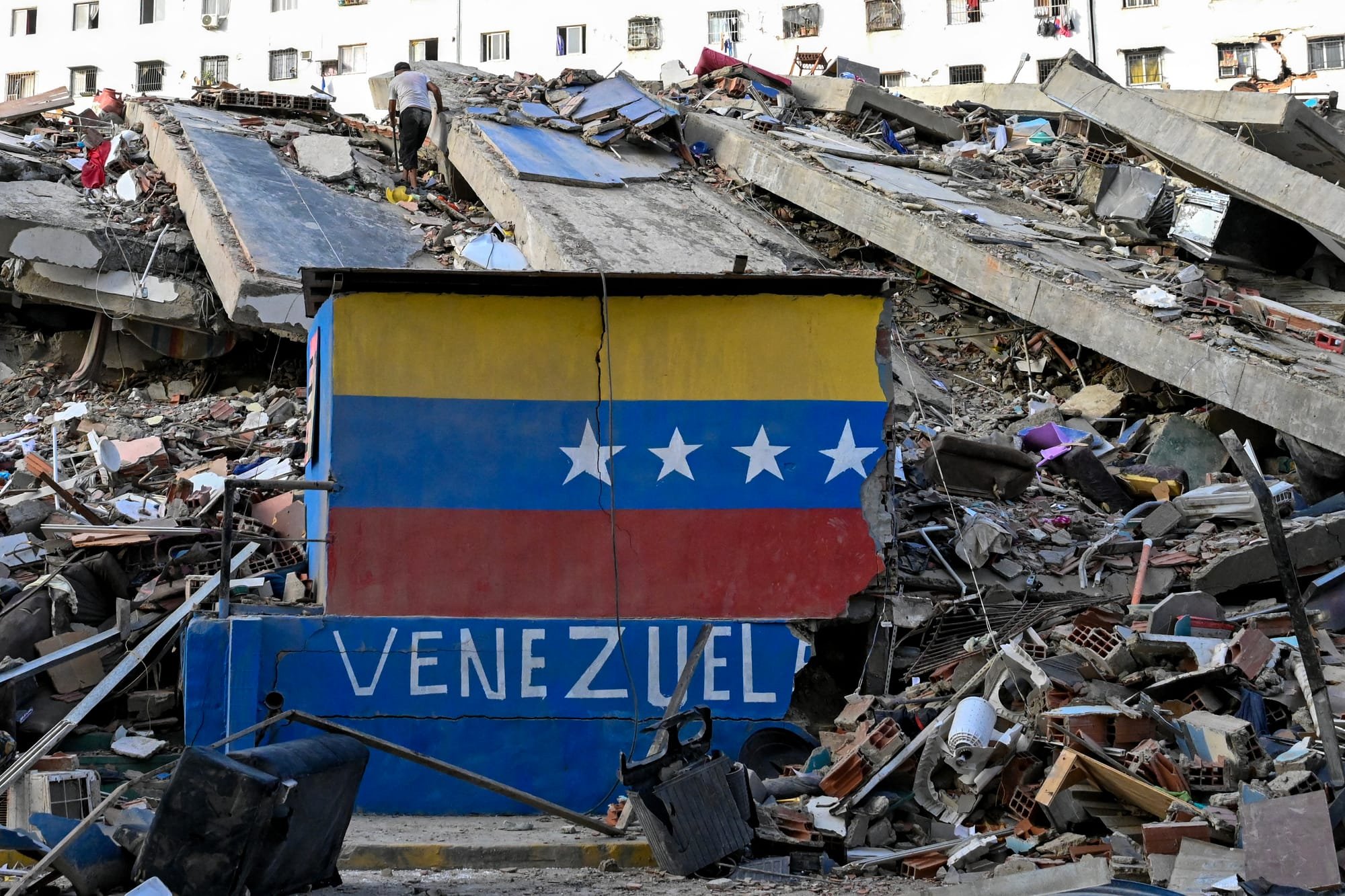 A partially destroyed building painted with the Venezuelan flag and "VENEZUELA" text sits amid concrete rubble, with a person walking across the debris-strewn upper level.